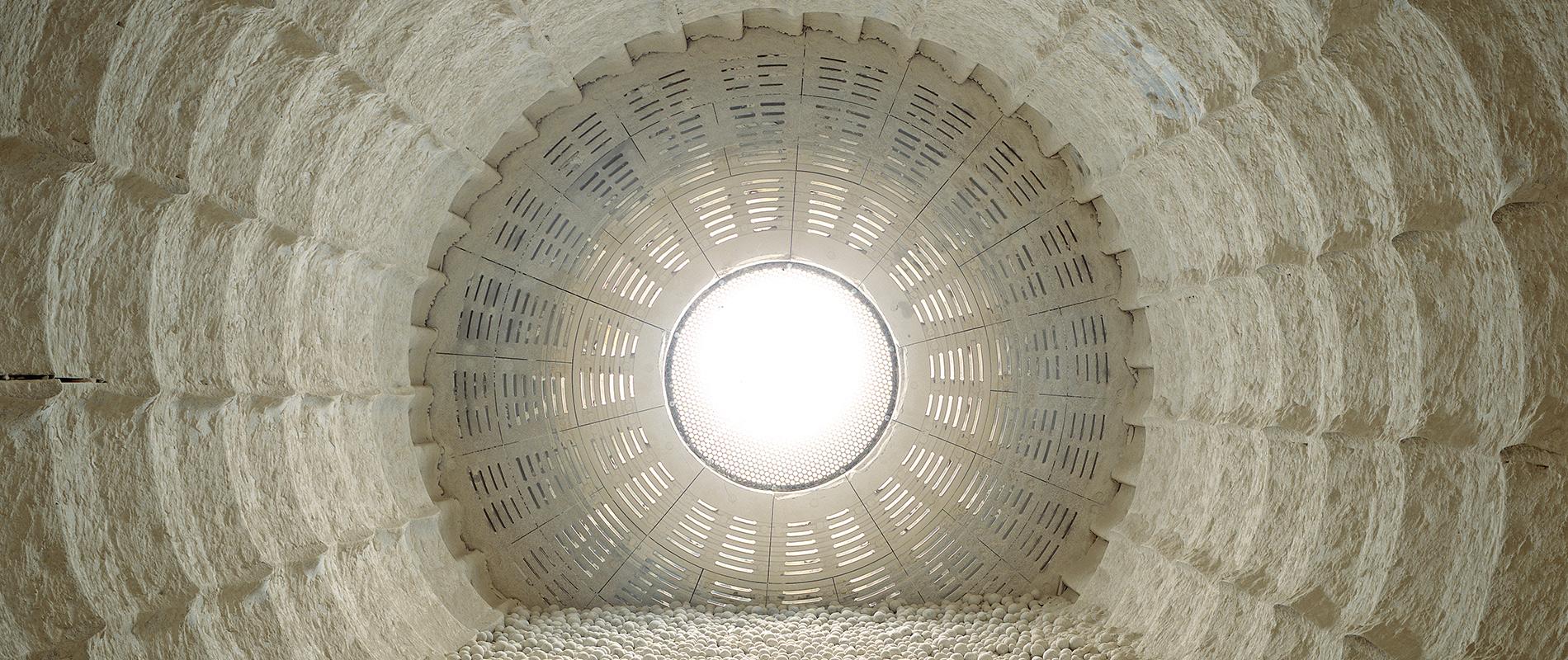 Blick von innen nach oben durch eine runde, strukturierte Öffnung, die von hell erleuchtetem Licht gefüllt ist. Die Wände umgeben den Betrachter mit einem Muster aus regelmäßig angeordneten, vertikalen und horizontalen Linien.