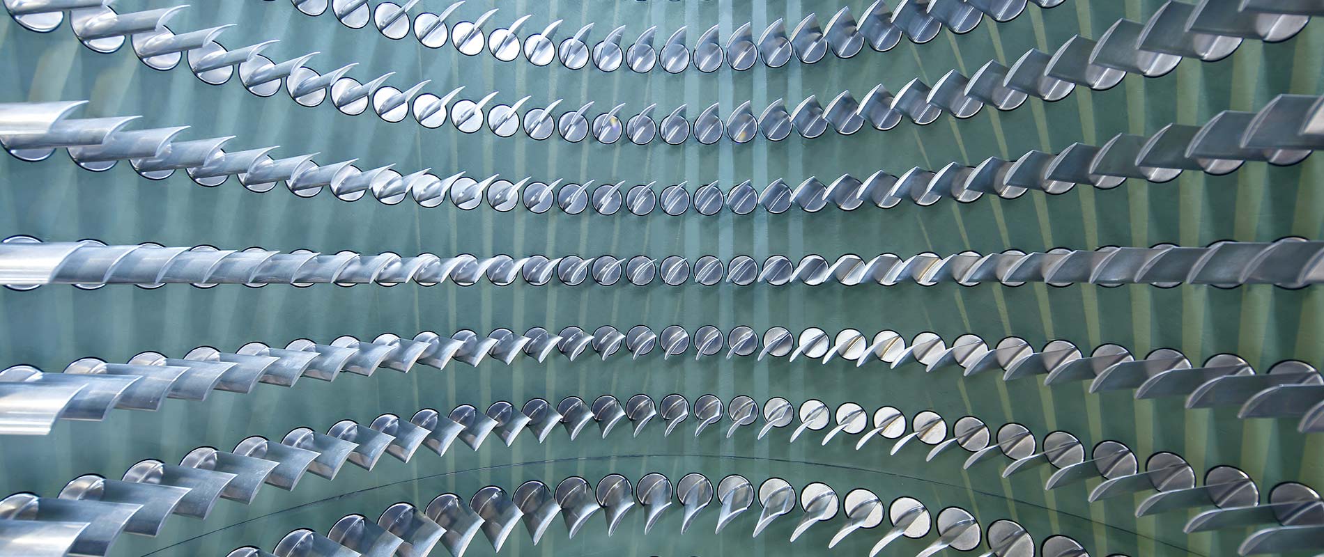 Eine Nahaufnahme von mehreren metallischen, gebogenen Objekten, die in gleichmäßigen Reihen angeordnet sind. Die Objekte leuchten silbern und sind in einer grünen, strukturierten Umgebung platziert, die eine moderne und futuristische Atmosphäre schafft.