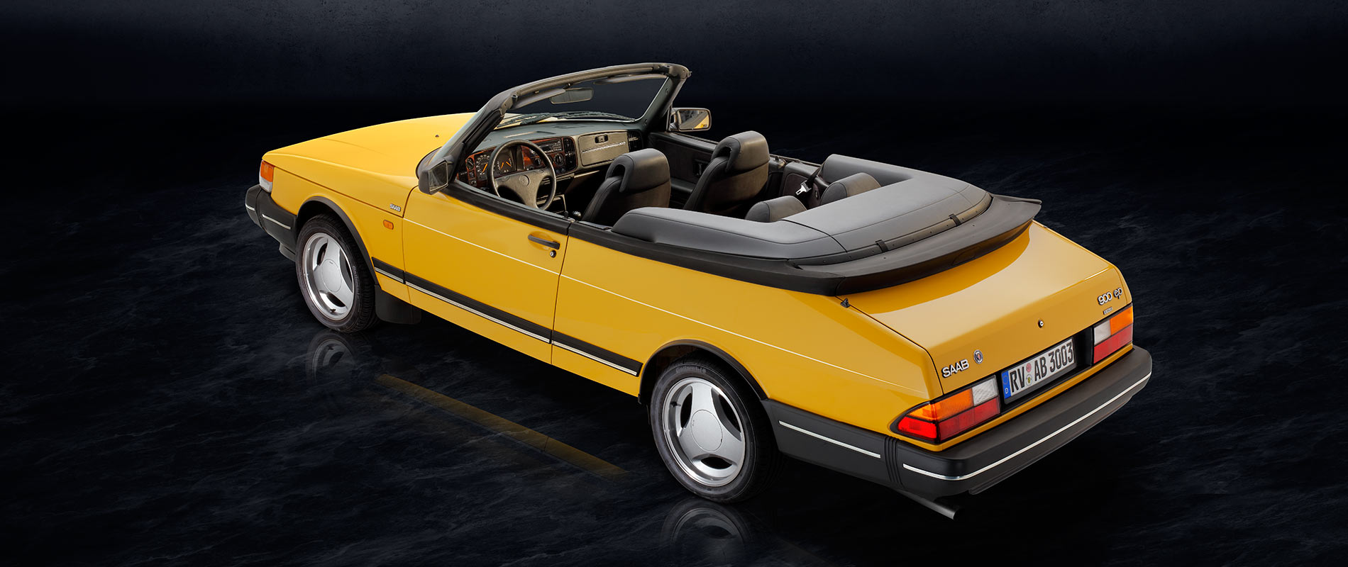 Ein gelbes Cabrio mit schwarzem Innenraum und gefaltetem Verdeck steht auf einer dunklen Oberfläche. Die Rückansicht zeigt die charakteristischen des Autos, einschließlich der Felgen und der Heckleuchten.